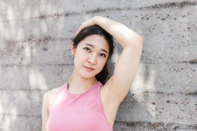 Portrait of a young woman posing with her hand on her head, leaning against a sunlit concrete wall.