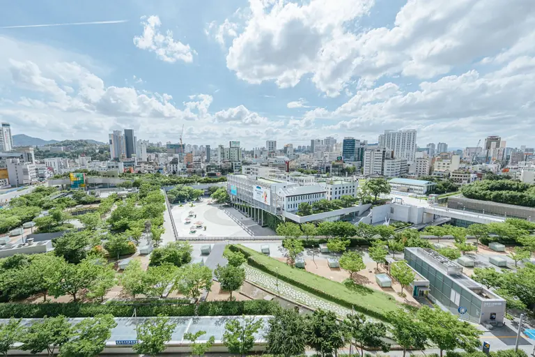 Aerial shot of a modern city with green parks, cloudy sky, and high-rise buildings.