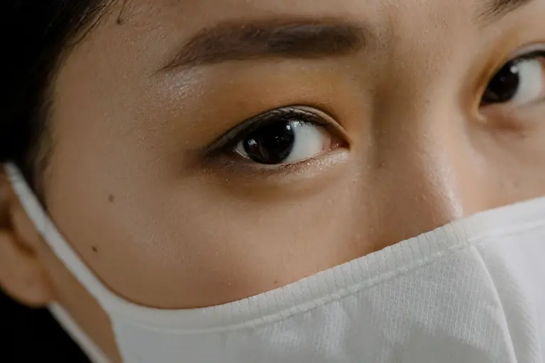 Close-up portrait of an Asian woman wearing a white face mask, highlighting eyes and eyebrows.