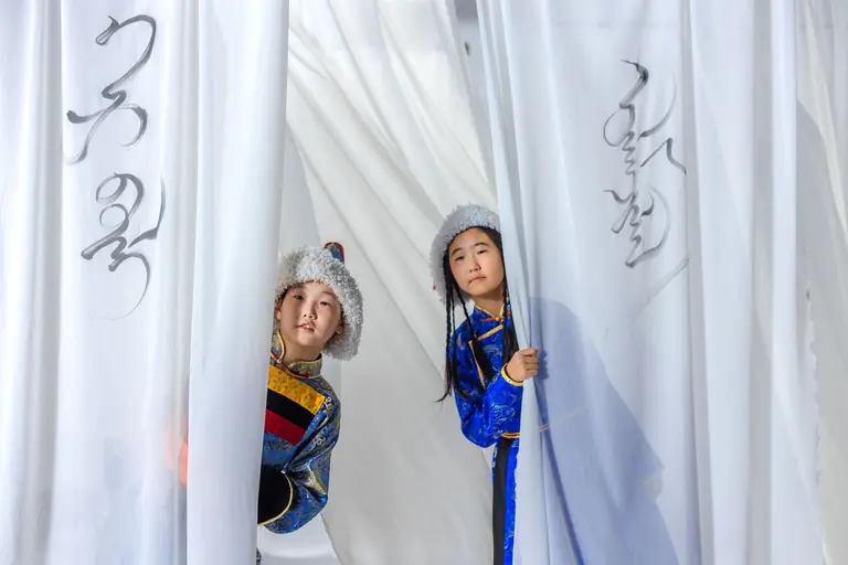 Two children in traditional attire peeking through white curtains, showcasing cultural elegance.