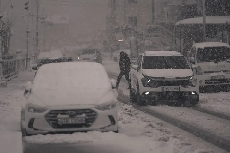 A snowy city street scene featuring vehicles and a person walking amidst heavy snowfall.