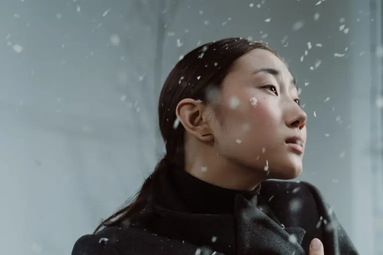 Asian woman in winter coat surrounded by snowflakes, looking thoughtful.