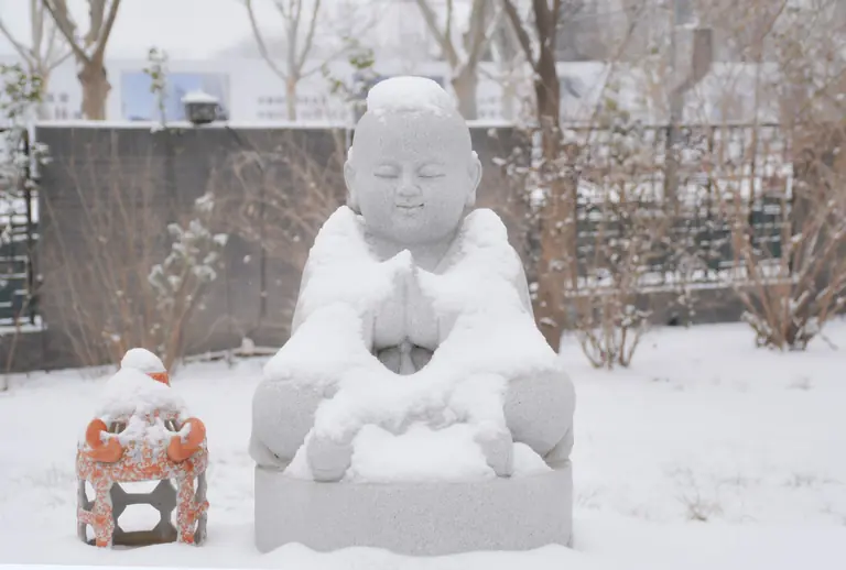A snow-covered Buddha statue meditating peacefully in a serene winter garden setting.