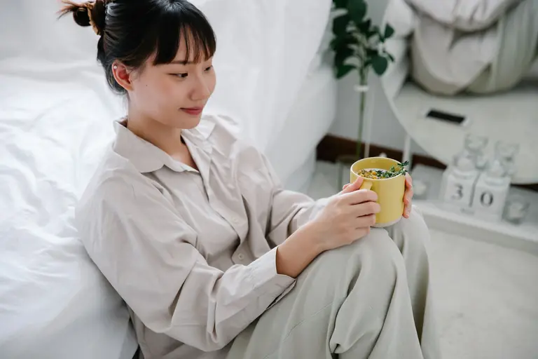 Young Asian woman in pajamas enjoying tea while relaxing on the bedroom floor.
