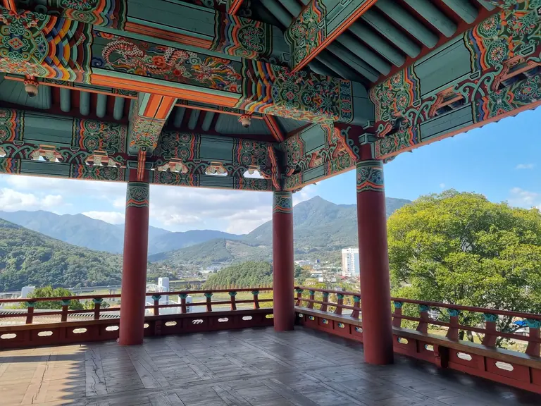 Colorful Korean pavilion overlooking mountains and cityscape under blue sky.
