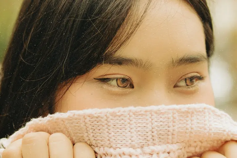 A young woman with captivating eyes gazes thoughtfully while covering her face with a pink sweater.