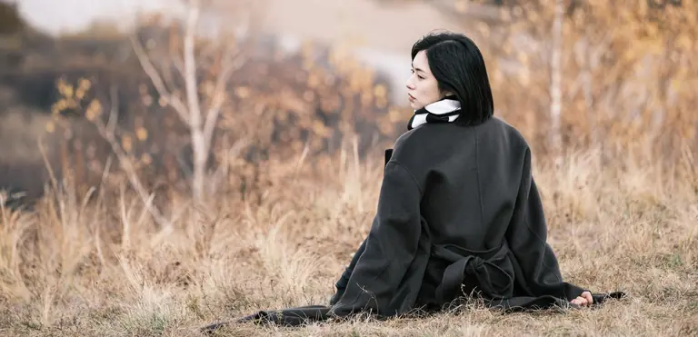 A woman in a black coat sits quietly in a meadow, surrounded by autumn scenery.