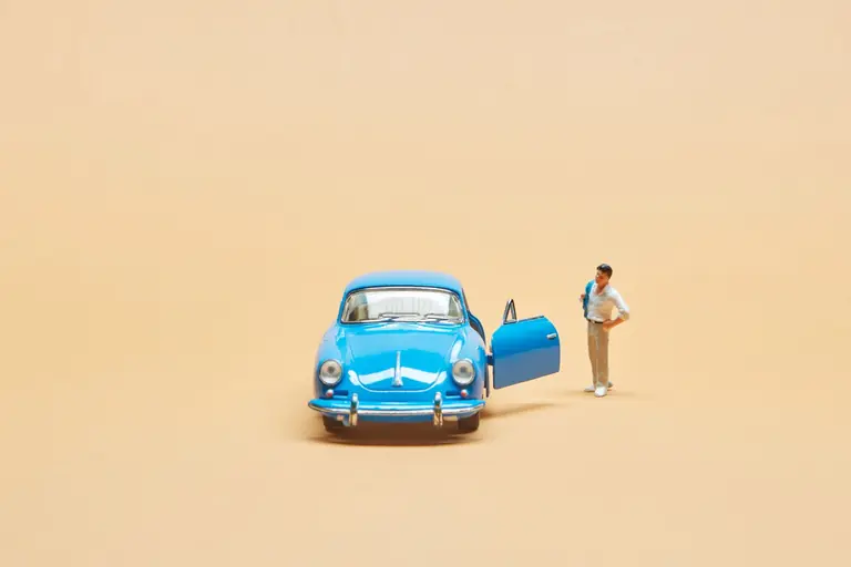 A miniature figurine stands beside a classic blue toy car on a beige background, creating a whimsical scene.