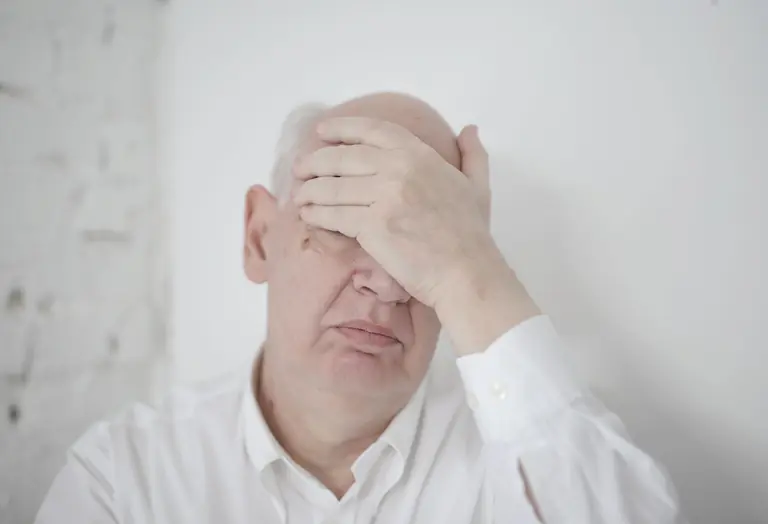 Disappointed senior bald man in white shirt covering face with hand while sitting near wall in light room