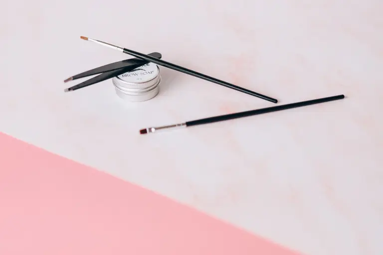 Aesthetic arrangement of makeup brushes and brow soap on a pastel pink background for beauty and fashion themes.