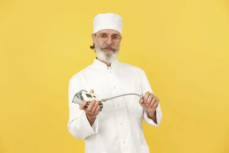 A senior chef in white attire holding a kitchen blender, standing against a yellow background.