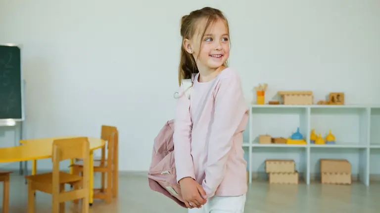 A joyful girl in a classroom setting, wearing a light pink shirt and holding a backpack.