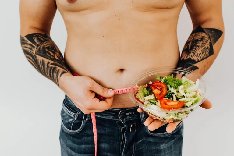 Tattooed man measuring waist while holding fresh salad, symbolizing healthy lifestyle and weight management.