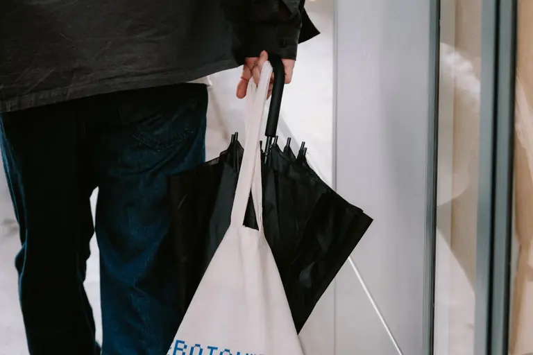 Man holding a black umbrella and canvas tote bag, walking on a city sidewalk.
