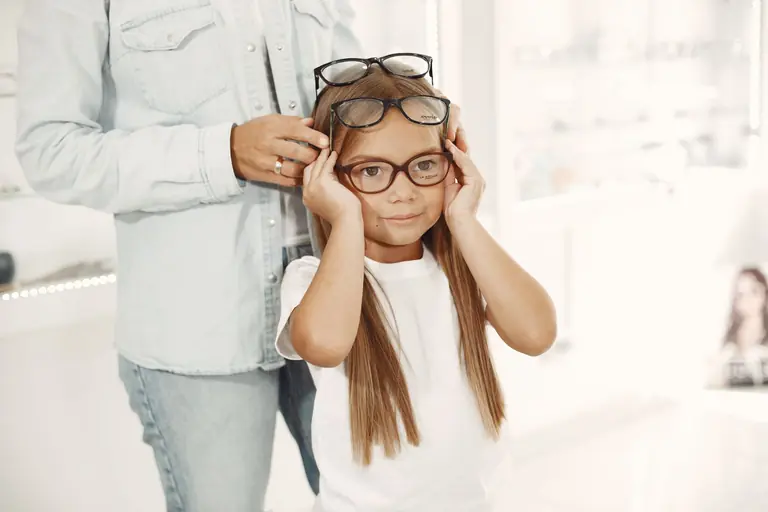 Young girl in a white shirt trying on glasses indoors, assisted by an adult.