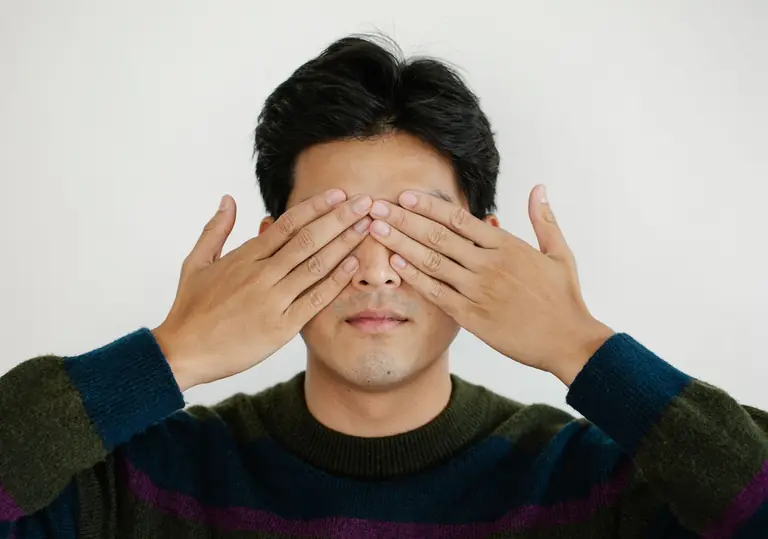 Close-up portrait of a man covering his eyes with his hands in a neutral background setting.