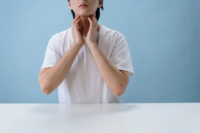 Close-up of an individual in a white crew neck t-shirt placing hands near neck, isolated on a blue backdrop.