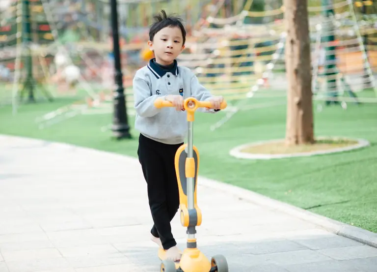A young boy enjoys a sunny day riding a scooter in a park with play structures.