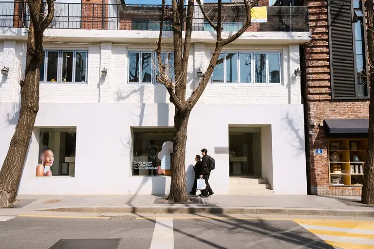 A city street scene showing a white building with trees casting shadows, and people walking.