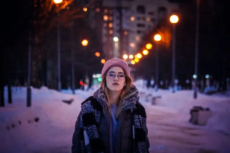 A stylish woman in winter clothing walks through an illuminated snowy park during the evening.