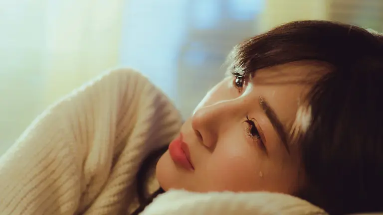 A woman in a sweater lying down and shedding a tear in a warm setting.