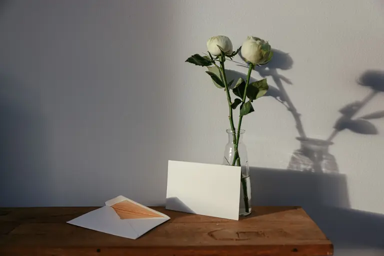 A minimalist scene showcasing white roses in a vase next to a blank card and envelope on a wooden surface.
