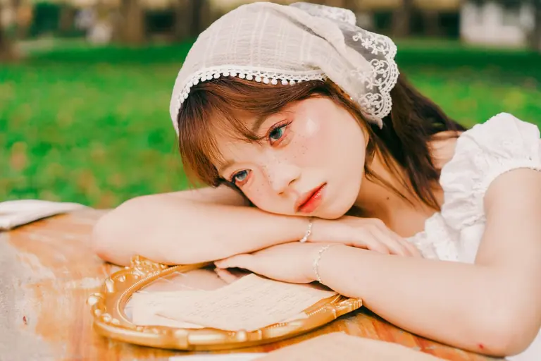 A young woman in a vintage dress with a lace headscarf rests her head on a table outdoors.