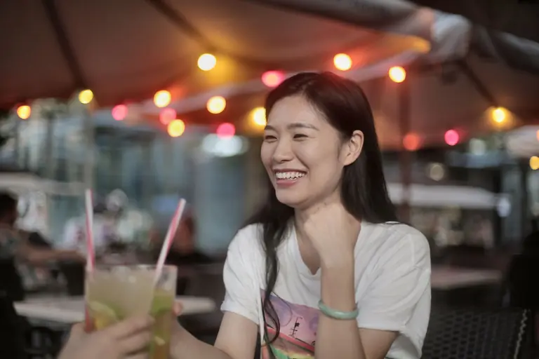 Asian woman smiling and enjoying cocktails at a lively outdoor bar with vibrant lighting.