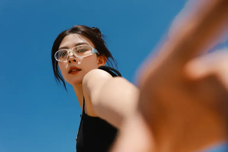 Stylish young woman in sunglasses reaches towards the camera with clear blue sky backdrop.
