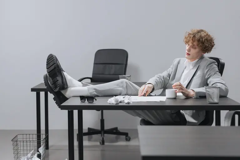 A curly-haired man in a suit relaxes with feet on desk, looking serious in a modern office setting.
